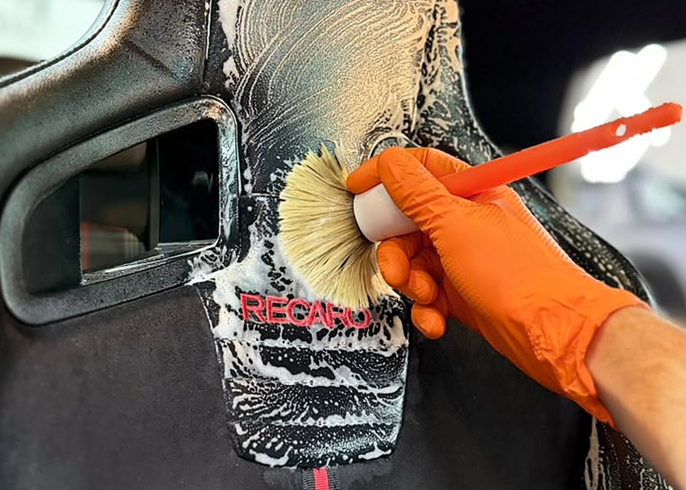 Escovação de banco Recaro com espuma de limpeza, usando escova e luva laranja — higienização interna profissional